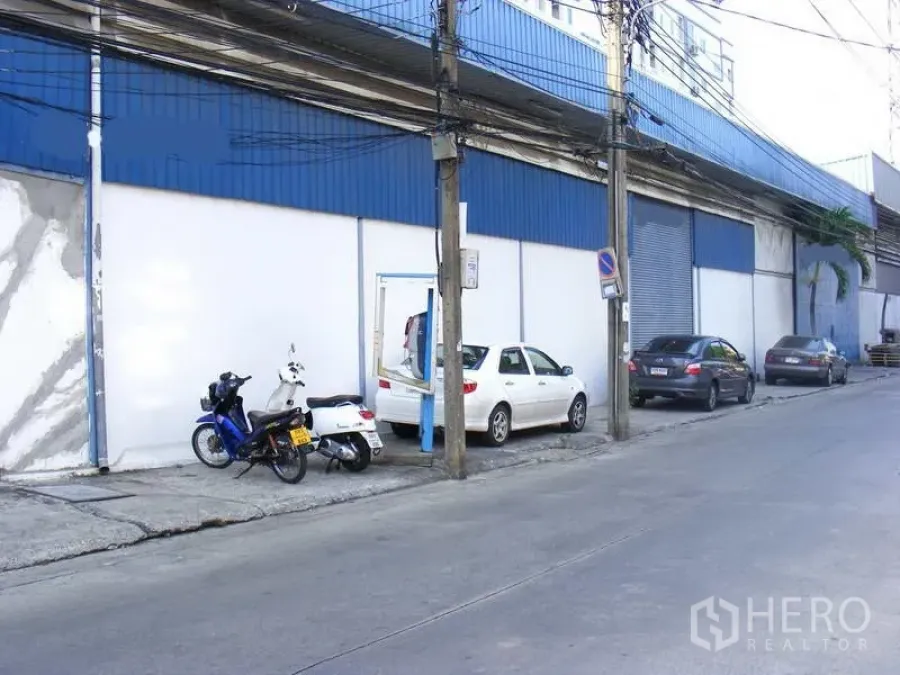 Warehouse for rent in Suan Luang, Bangkok - Street-facing warehouse exterior in Suan Luang, Bangkok with blue cladding, white walls, roller shutter door and roadside parking for cars and motorbikes.