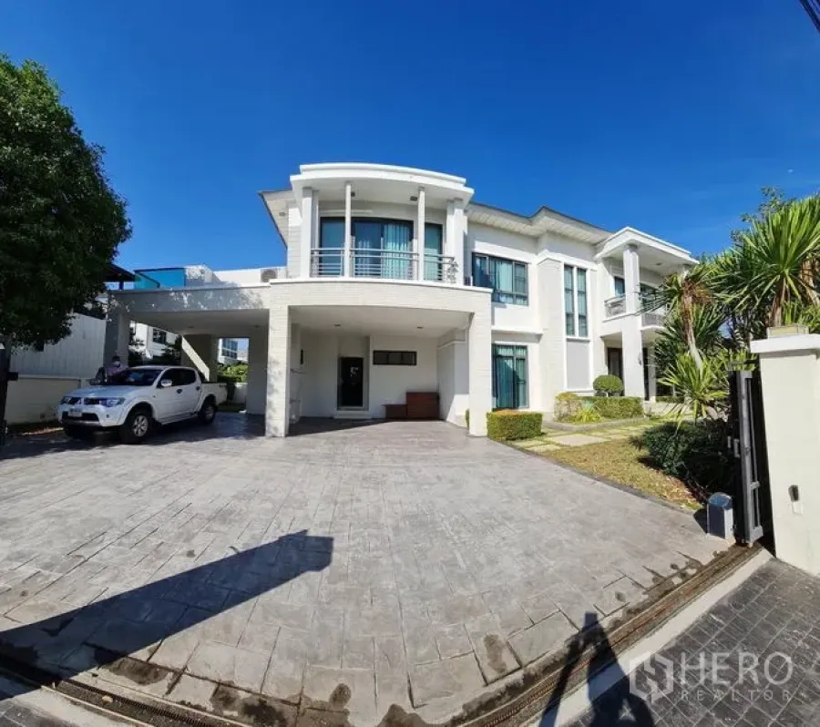 Detached House for sale in Khlong Toei, Bangkok - Modern two-storey white detached house with curved balcony, large paved driveway, carport and landscaped garden under blue sky.