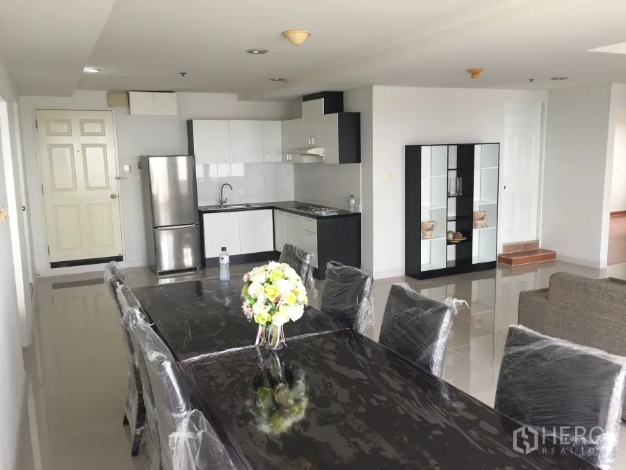 Condominium for rent in Khlong Toei, Bangkok - Open-plan dining area with black table and chairs beside a white L-shaped kitchen with a stainless-steel fridge.
