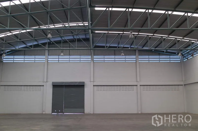 Factory for rent in Pathum Thani - High-ceiling factory warehouse interior with steel truss roof, clerestory vents and a wide roller shutter door.