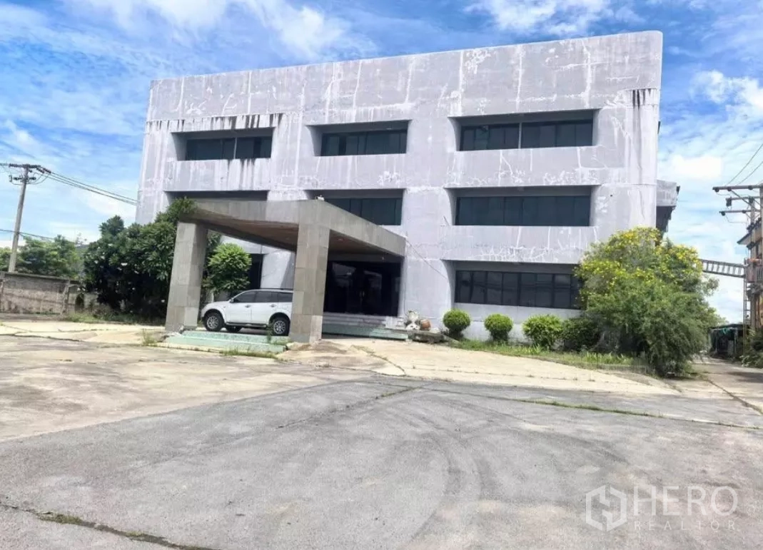 Factory for sale in Bang Phli, Samut Prakan - Front facade of the three-storey industrial office with porte-cochère and landscaped shrubs.