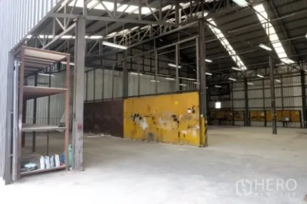 Warehouse for rent in Pak Kret, Nonthaburi - Spacious warehouse interior with high steel trusses, concrete floor, and a yellow partition wall.
