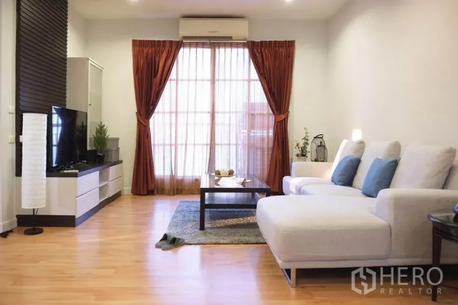 Condominium for rent in Khlong Toei, Bangkok - Spacious living area with L-shaped sofa, coffee table, wood flooring, and balcony-style doors.