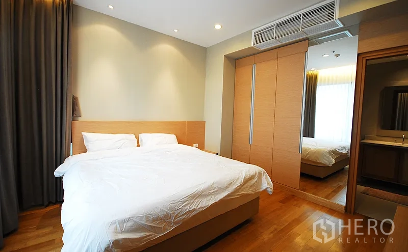 Condominium for rent in Khlong Toei, Bangkok - Spacious bedroom with king bed, mirrored built-in wardrobe, wood floors and doorway to modern bathroom.