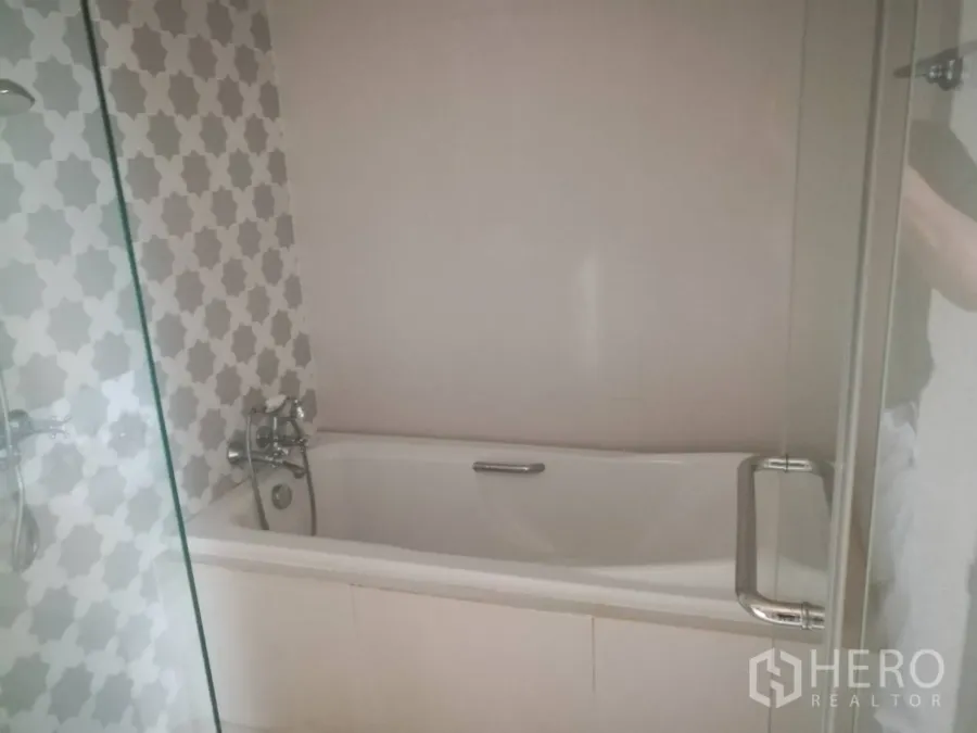 Condominium for sale in Khlong Toei, Bangkok - Modern bathroom with white bathtub, patterned gray tiles and a glass shower screen.