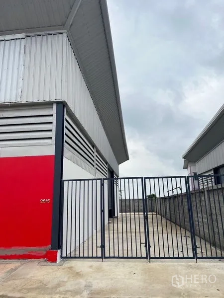 Warehouse for rent or sale in Sai Noi, Nonthaburi - Corner view of metal-clad warehouse with red accent wall and gated driveway.