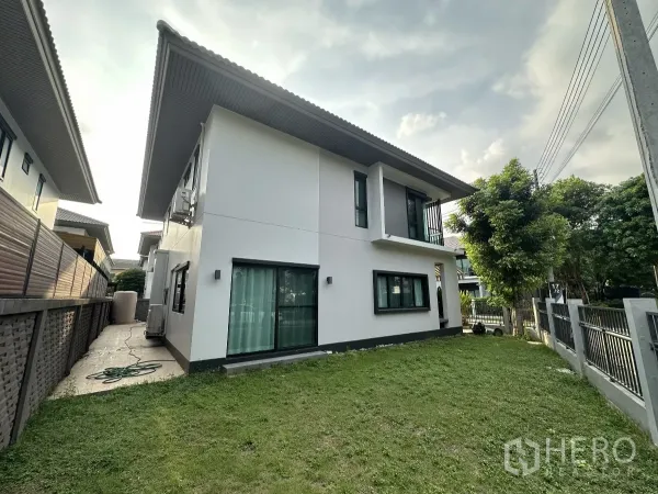 Detached House for rent or sale in Lam Luk Ka, Pathum Thani - Side view of the house with a green lawn, sliding glass door and perimeter fence shaded by mature trees.
