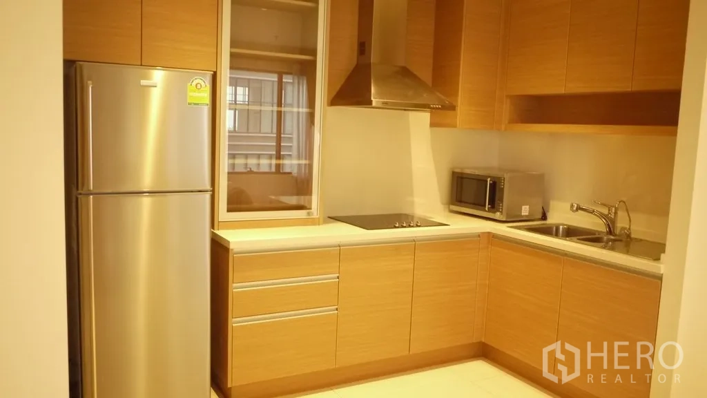 Condominium for rent in Khlong Toei, Bangkok - Modern L-shaped kitchen with wood cabinets, electric cooktop, range hood, stainless refrigerator, microwave and sink.