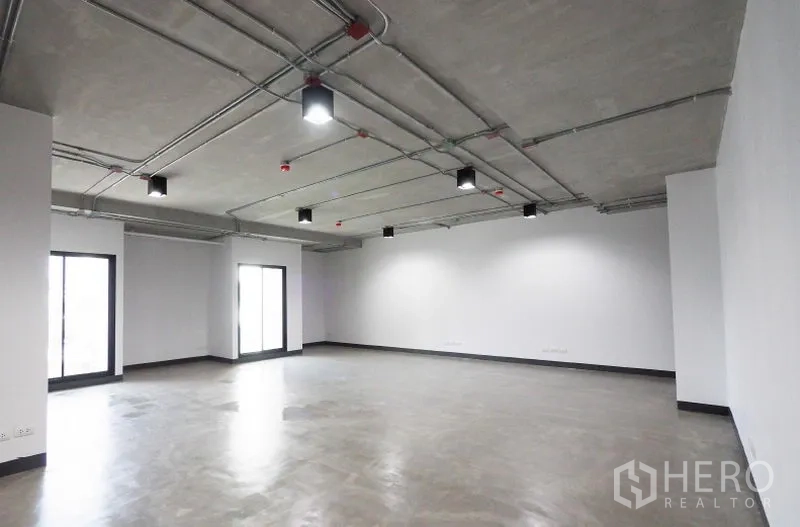 Office Space for rent in Khlong Toei, Bangkok - Spacious corner office with large windows, visible conduits on a concrete ceiling, LED lights, white walls and smooth grey flooring.