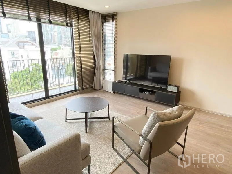 Condominium for rent in Pathum Wan, Bangkok - Living room with sofa, armchair, TV console and wide balcony showcasing city and greenery views.