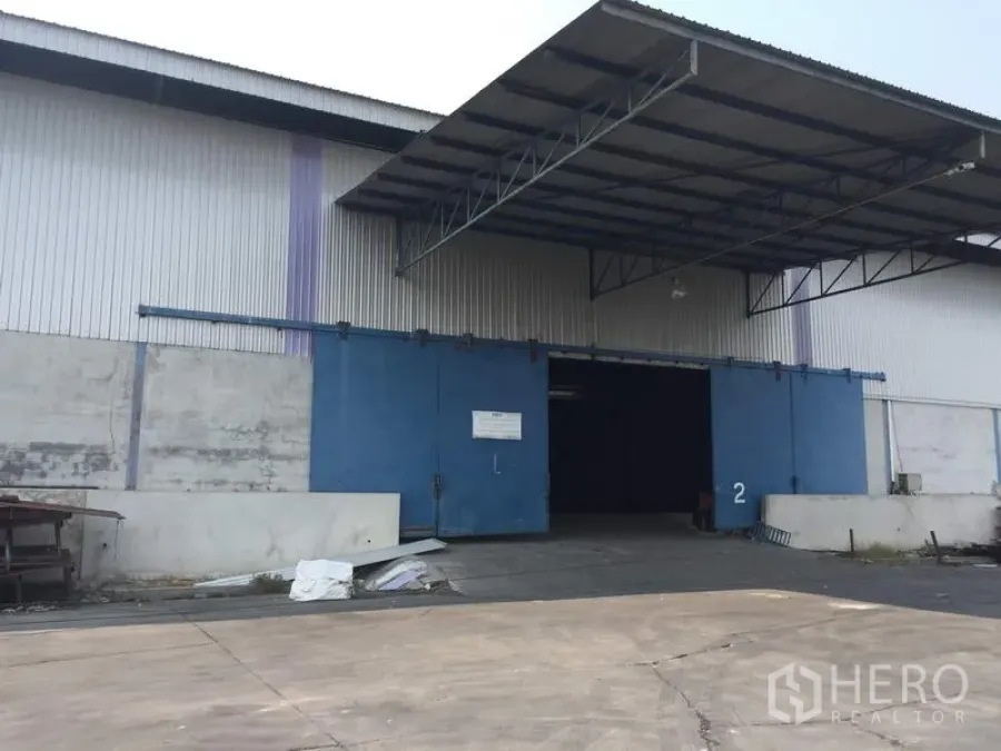 货栈 出租在Pathum Thani - 仓库外立面，蓝色大型滑动门及带雨棚的装卸区。