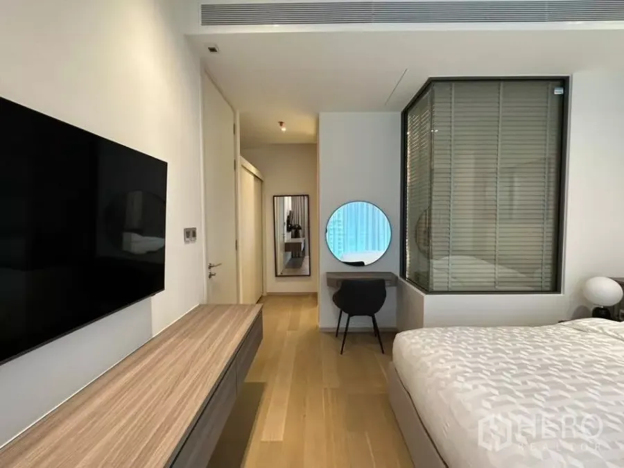 Condominium for rent in Bangkok - Bright Thonglor condo bedroom with wood floors, wall-mounted TV, study desk with round mirror, and glass partition with blinds.