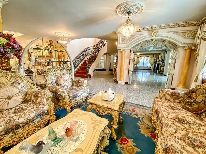 Detached House for rent or sale in Bang Khen, Bangkok - Spacious lounge with classical sofas, arched doorway, and curved staircase with wrought-iron rails.