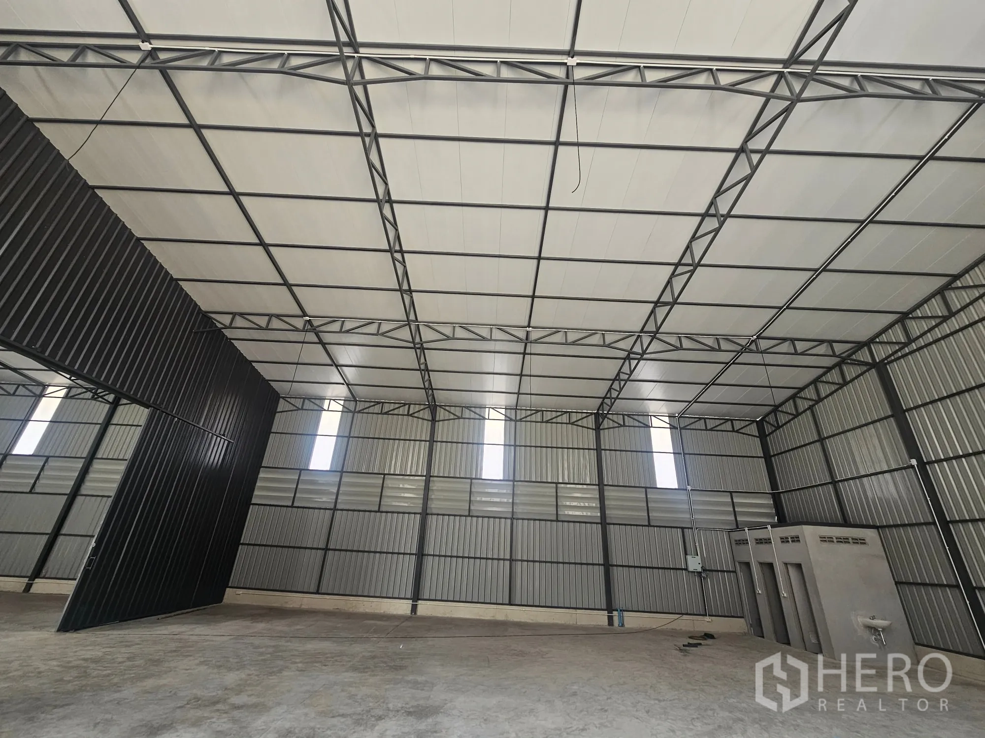 Warehouse for rent in Bang Khen, Bangkok - Steel-frame warehouse interior with panelled ceiling, tall sliding door, and a small restroom block along the wall.