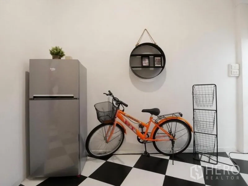 Townhouse for rent in Khlong Toei, Bangkok - Utility nook with a silver refrigerator, orange bicycle, round wall shelf and wire storage rack.