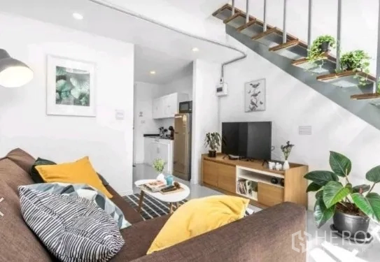 Townhouse for rent in Khlong Toei, Bangkok - Cozy living area with TV console, indoor plants, and a kitchen tucked beneath the staircase.