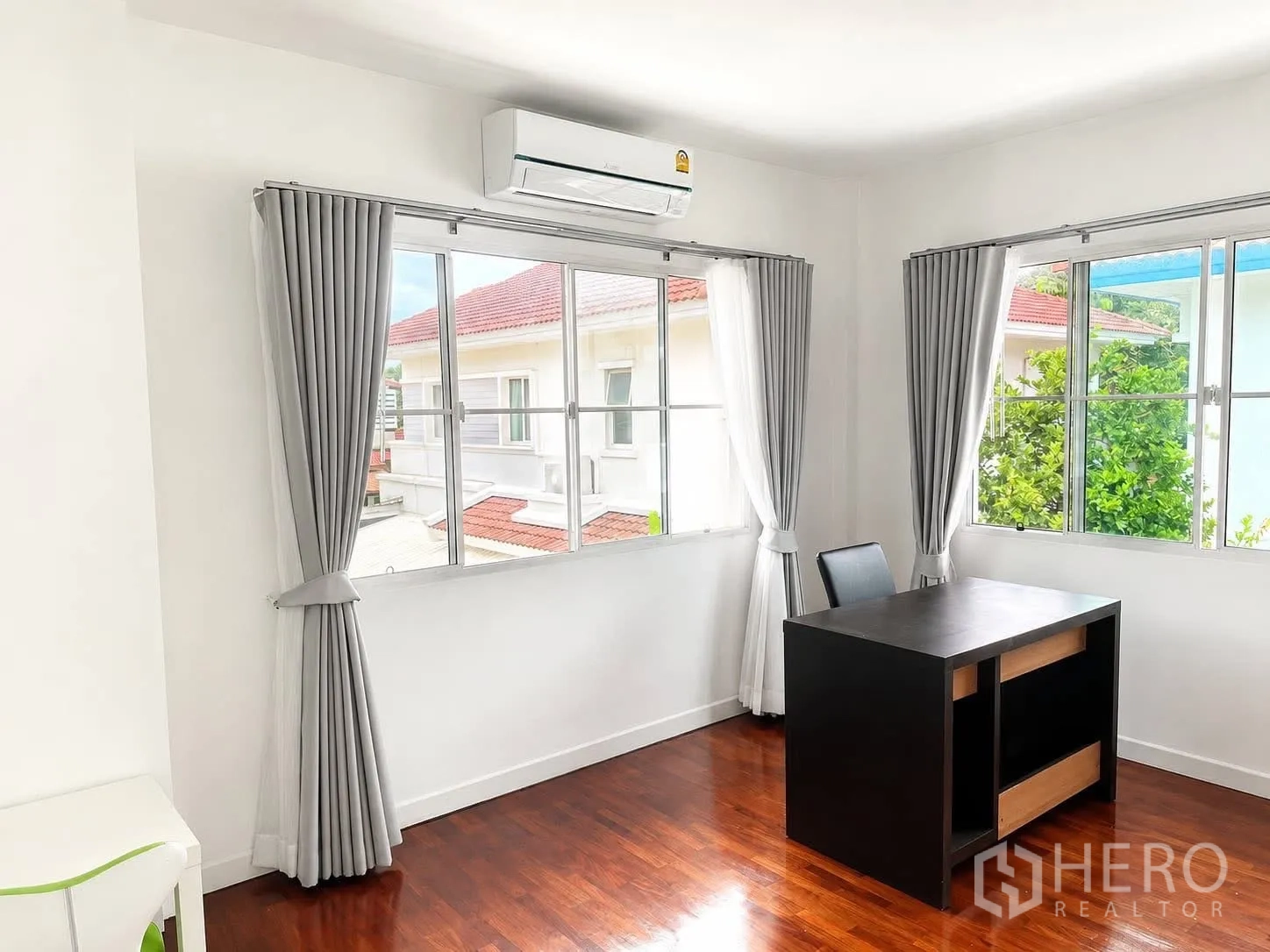 Detached House for rent in Prawet, Bangkok - Bright study corner with large windows, gray curtains, air conditioner, and a black desk on wood flooring.