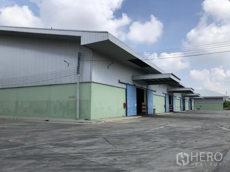 Warehouse for rent in Phra Nakhon Si Ayutthaya - Row of warehouse units with blue sliding doors, loading canopies and an expansive concrete yard.