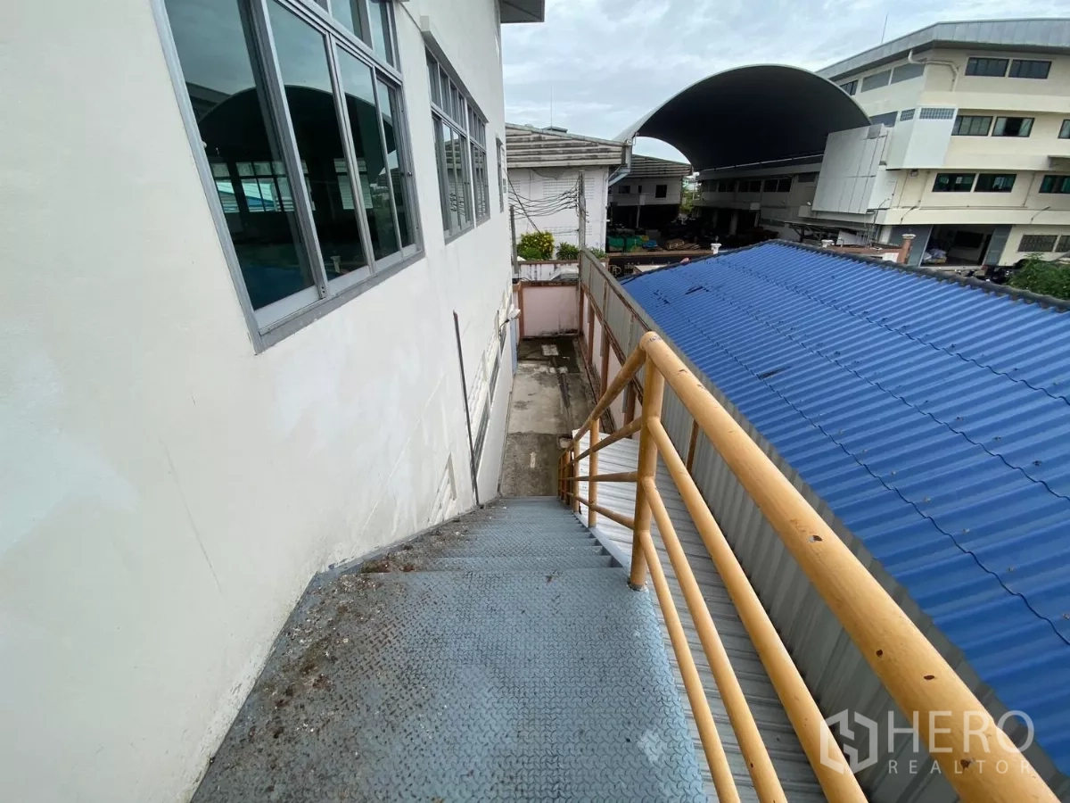 Factory for rent in Khlong Toei, Bangkok - Exterior metal staircase with yellow handrail beside the factory wall and a blue roof.