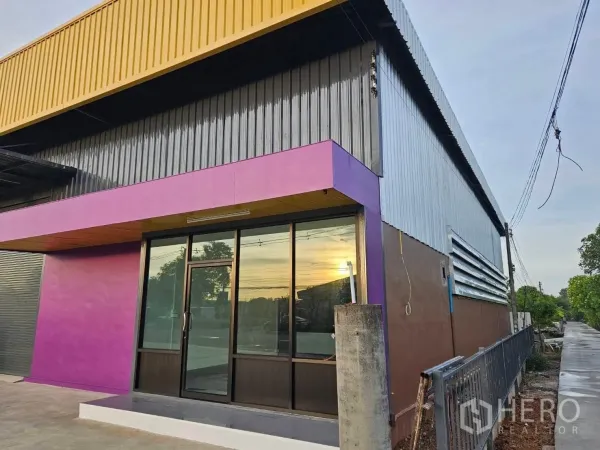 Warehouse for rent in Lam Luk Ka, Pathum Thani - Corner facade showing the glass-front office entrance with a purple accent beneath yellow roof panels.