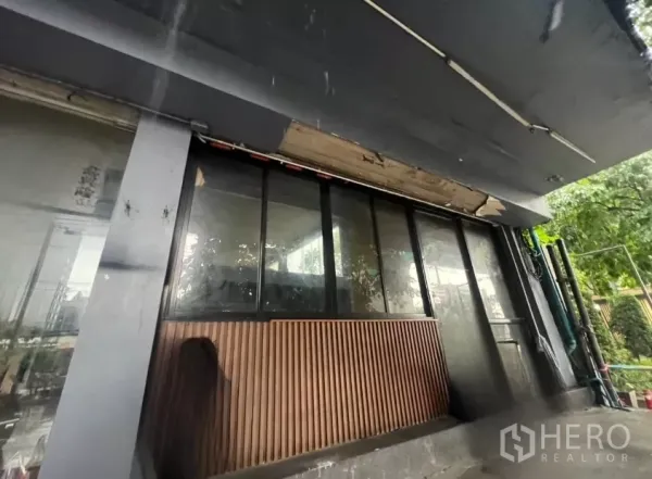 Shophouse for rent in Khlong Toei, Bangkok - Street-facing shopfront with black awning, large glass panels and vertical wood cladding beside roadside greenery.