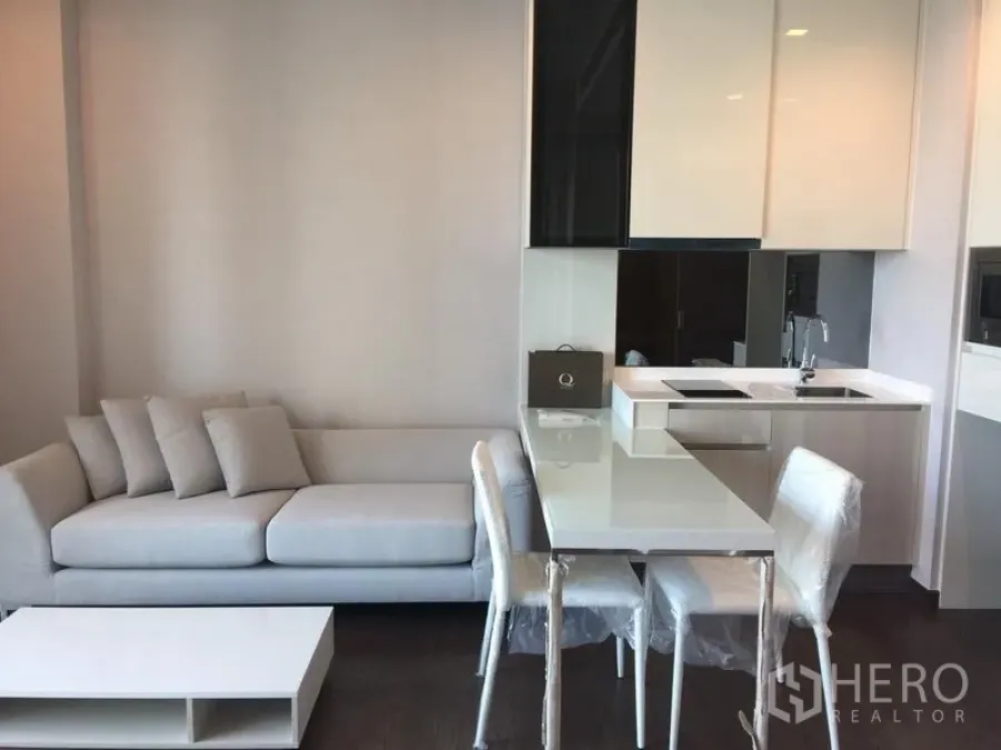 Condominium for rent in Watthana, Bangkok - Open-plan living area with light gray sofa, dining set, and kitchenette with induction cooktop and sink.