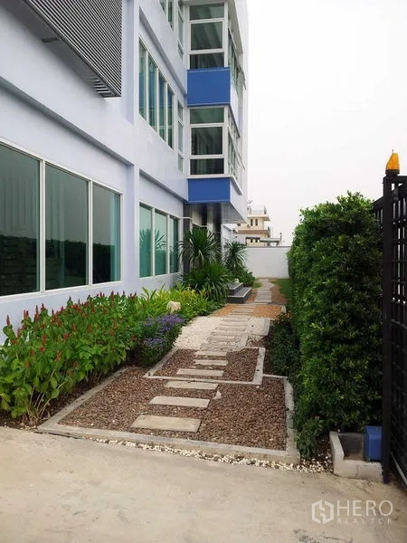 Warehouse for rent or sale in Bang Phli, Samut Prakan - Side garden with stepping-stone path, shrubs, and windows along the warehouse exterior.