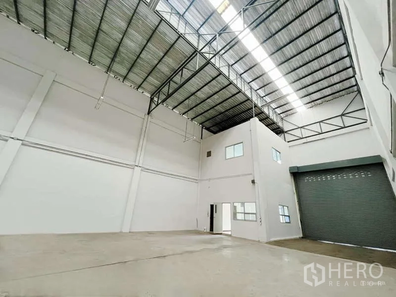 Factory for rent in Mueang Samut Sakhon, Samut Sakhon - Spacious high-ceiling factory interior with skylight, steel truss roof, and roller shutter beside an internal office block.
