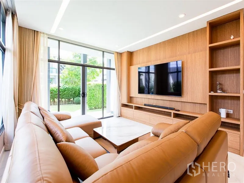 Detached House for rent or sale in Prawet, Bangkok - Living room with leather sectional, TV wall and views to the garden through sliding doors.
