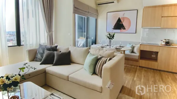 Condominium for rent or sale in Khlong Toei, Bangkok - Living area with corner sofa, tall windows, and adjacent dining nook and kitchen.
