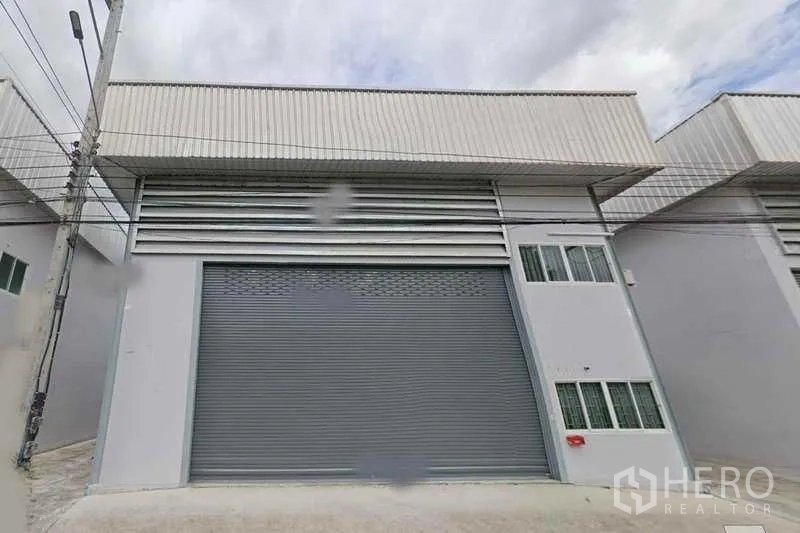 Warehouse for rent in Thanyaburi, Pathum Thani - Modern light-gray warehouse exterior in Thanyaburi with a wide metal roller shutter, louvered vents, twin windows and a concrete loading apron under a cloudy sky.