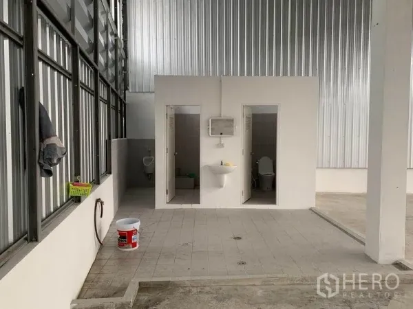 Warehouse for rent in Mueang Pathum Thani, Pathum Thani - On-site restroom block inside the warehouse with toilets and a handwash basin against metal wall cladding.