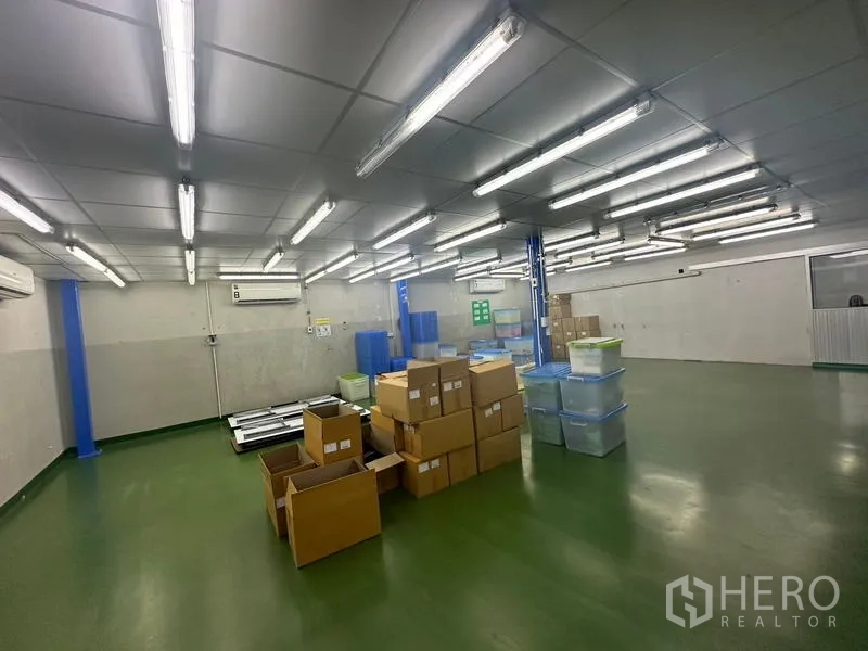 Warehouse for rent or sale in Lam Luk Ka, Pathum Thani - Large fluorescent‑lit storage area with boxes and plastic bins on a green floor.