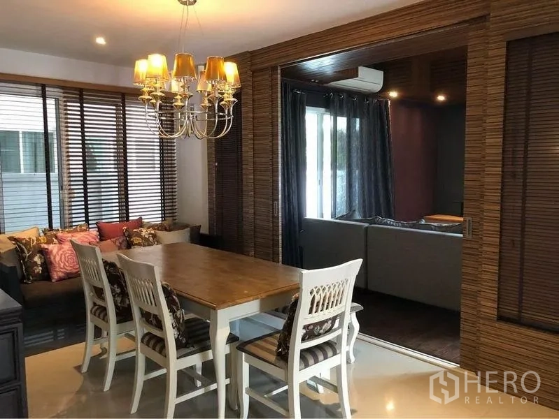 Detached House for rent or sale in Prawet, Bangkok - Dining area with wooden table, chandelier, and wide windows with blinds.