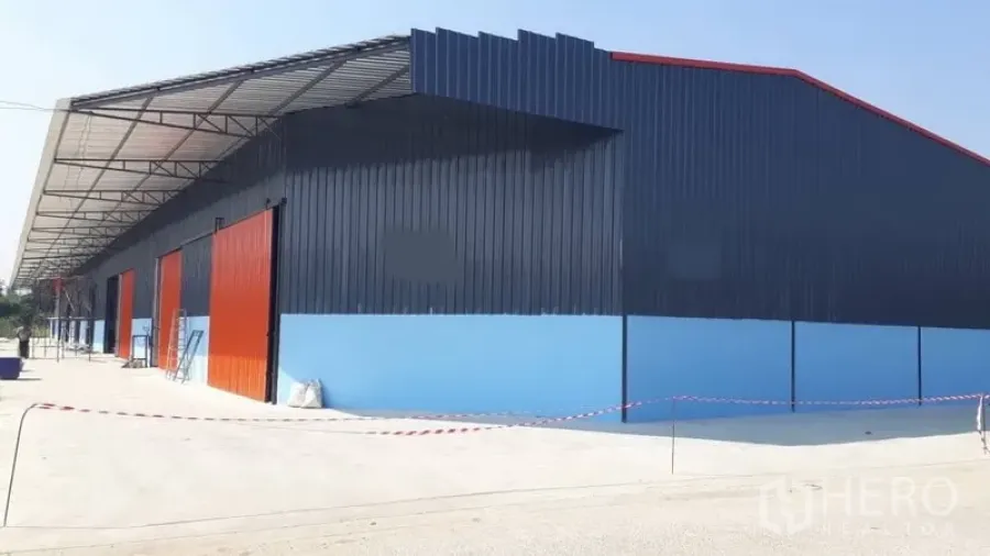Warehouse for rent in Pak Kret, Nonthaburi - Modern blue steel-clad warehouse in Pak Kret with red sliding doors and a wide concrete yard under a long loading canopy.