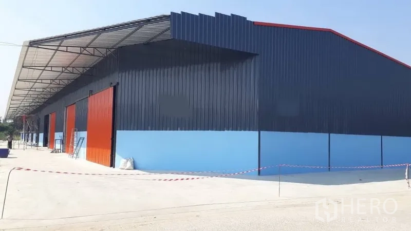 Warehouse for rent in Pak Kret, Nonthaburi - Modern blue steel-clad warehouse in Pak Kret with red sliding doors and a wide concrete yard under a long loading canopy.
