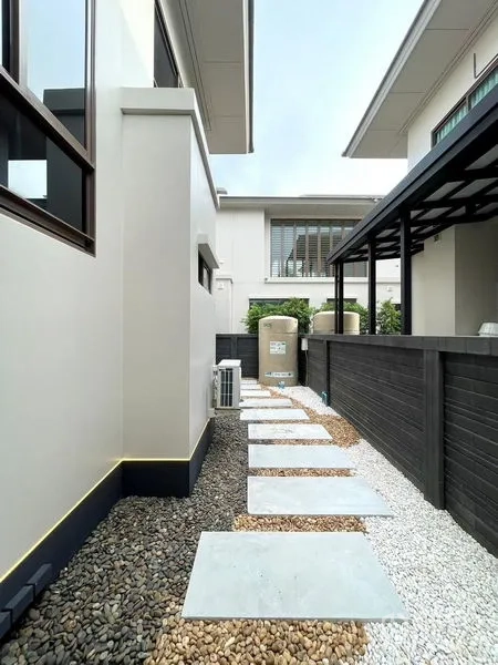 Detached House for sale in Bang Kapi, Bangkok - Side yard with stepping-stone path over pebbles, utility tanks and air-conditioning units along a fence.