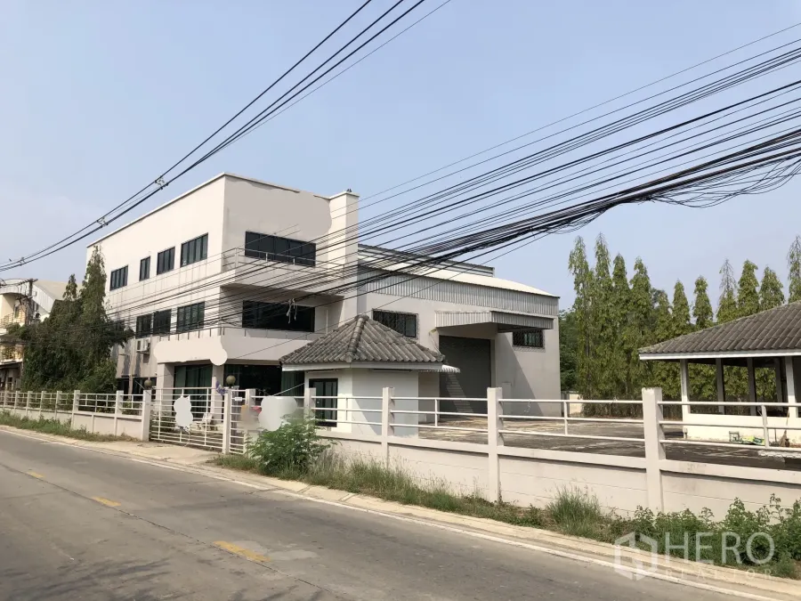 Factory for sale in Lam Luk Ka, Pathum Thani - Front view of a modern factory with office wing, loading bay and gated perimeter along a quiet road.