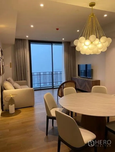 Condominium for rent in Pathum Wan, Bangkok - Cozy lounge with round dining set, statement pendant light, TV cabinet, and floor-to-ceiling glass opening to a balcony.