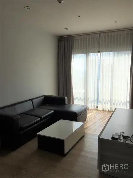 Condominium for rent in Bangkok - Bright living room with black leather L-shaped sofa, coffee table and floor-to-ceiling sheer curtains.