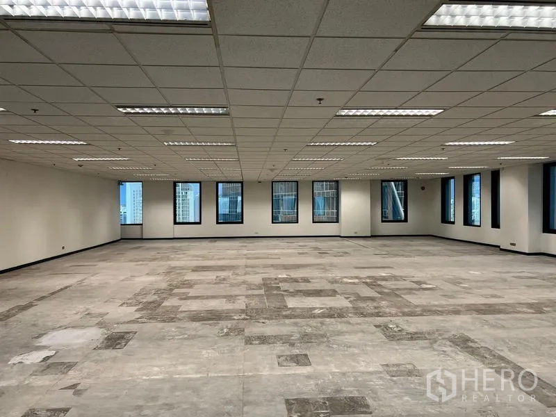 Office Space for rent in Khlong Toei, Bangkok - Open office area with patchwork flooring and many windows overlooking Bangkok skyscrapers beneath a grid ceiling.
