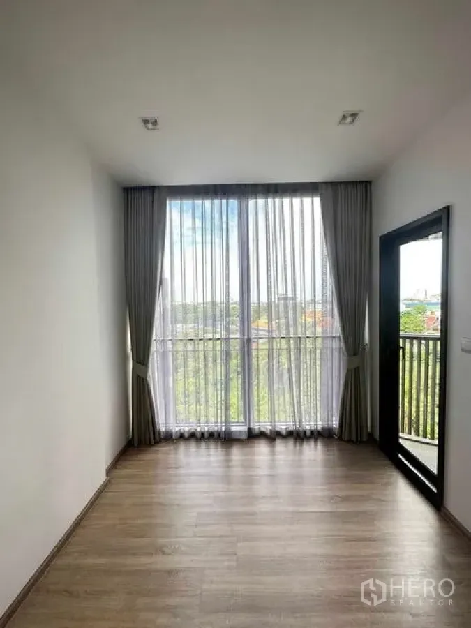 Condominium for sale in Watthana, Bangkok - Room with full‑height window and balcony door, sheer curtains, and treetop outlook.