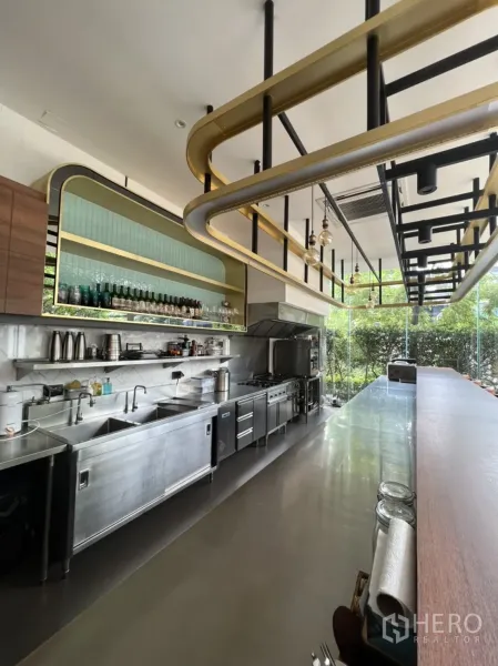 Retail Space for rent in Watthana, Bangkok - Full stainless commercial kitchen line with sinks, ovens and shelves beneath a gold feature rack facing garden views.