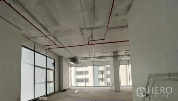 Retail Space for rent in Khlong Toei, Bangkok - Open-plan shell retail unit with high ceiling, exposed red sprinkler pipes and floor-to-ceiling windows overlooking city buildings in Khlong Toei.