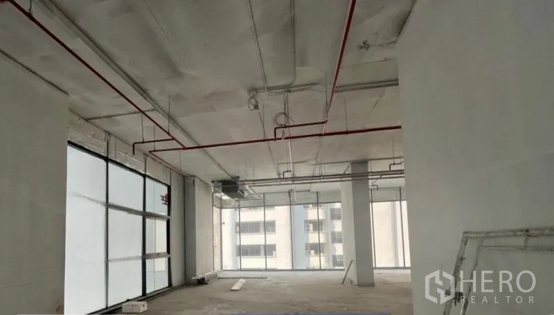Retail Space for rent in Khlong Toei, Bangkok - Open-plan shell retail unit with high ceiling, exposed red sprinkler pipes and floor-to-ceiling windows overlooking city buildings in Khlong Toei.