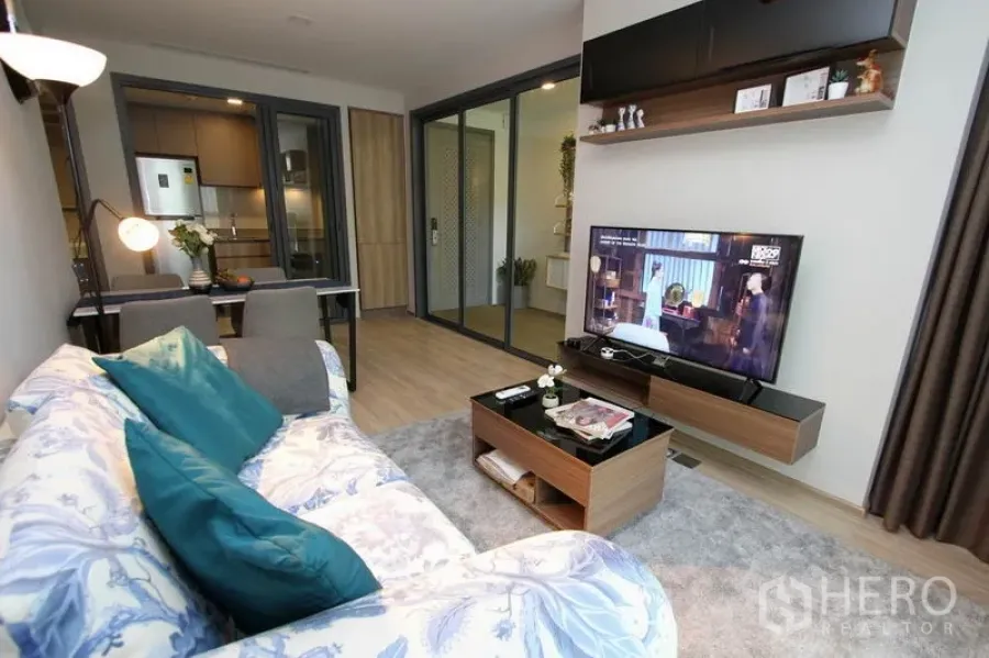 Condominium for rent or sale in Watthana, Bangkok - Bright Ekkamai condo living room with floral sofa, teal cushions, wall-mounted TV, wood floors and sliding glass doors to the kitchen.