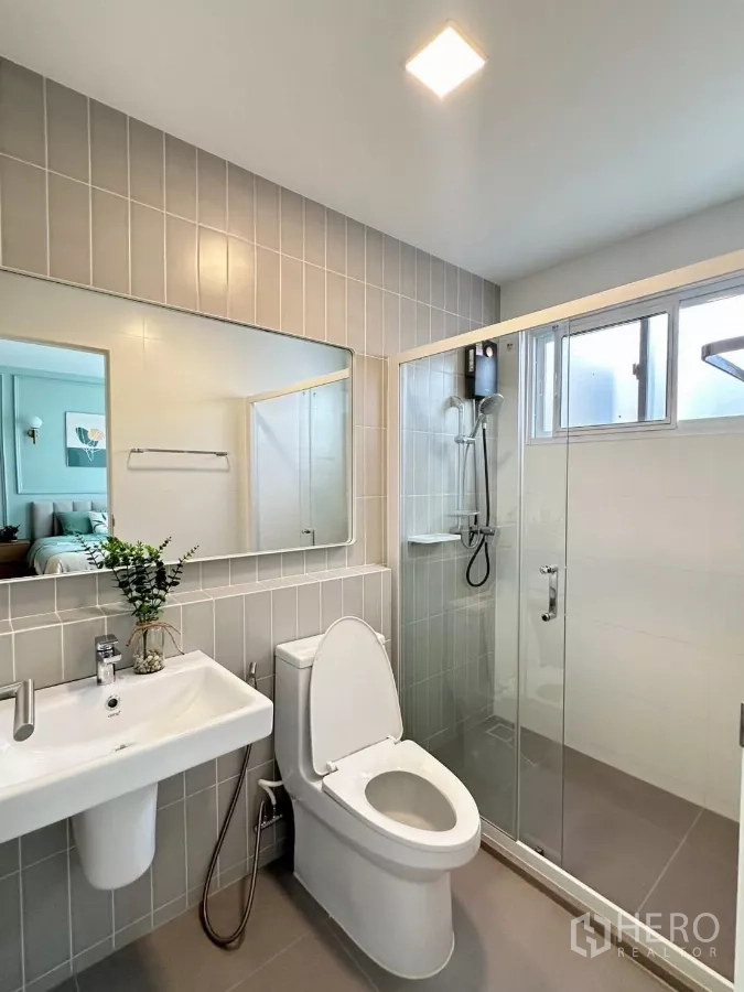 Townhouse for rent in Prawet, Bangkok - Bathroom with large mirror, glass-enclosed shower and gray tiles.