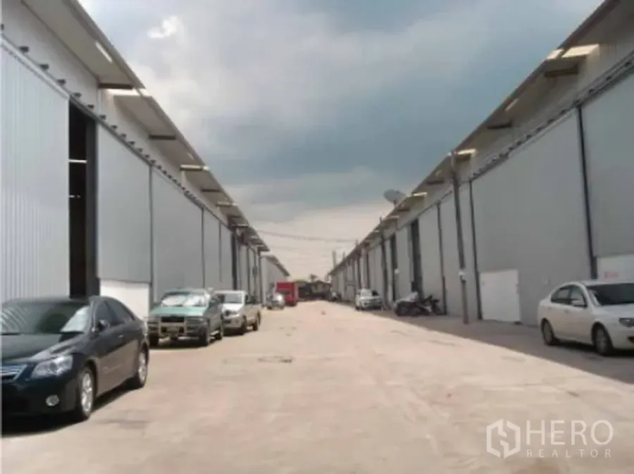 Warehouse for rent in Phra Pradaeng, Samut Prakan - Row of modern gray warehouse buildings with a wide internal road, street lighting and parked vehicles.