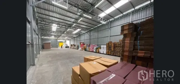 Warehouse for rent in Don Mueang, Bangkok - High-clearance metal warehouse interior with skylights, wide aisle and stacked cartons for storage.