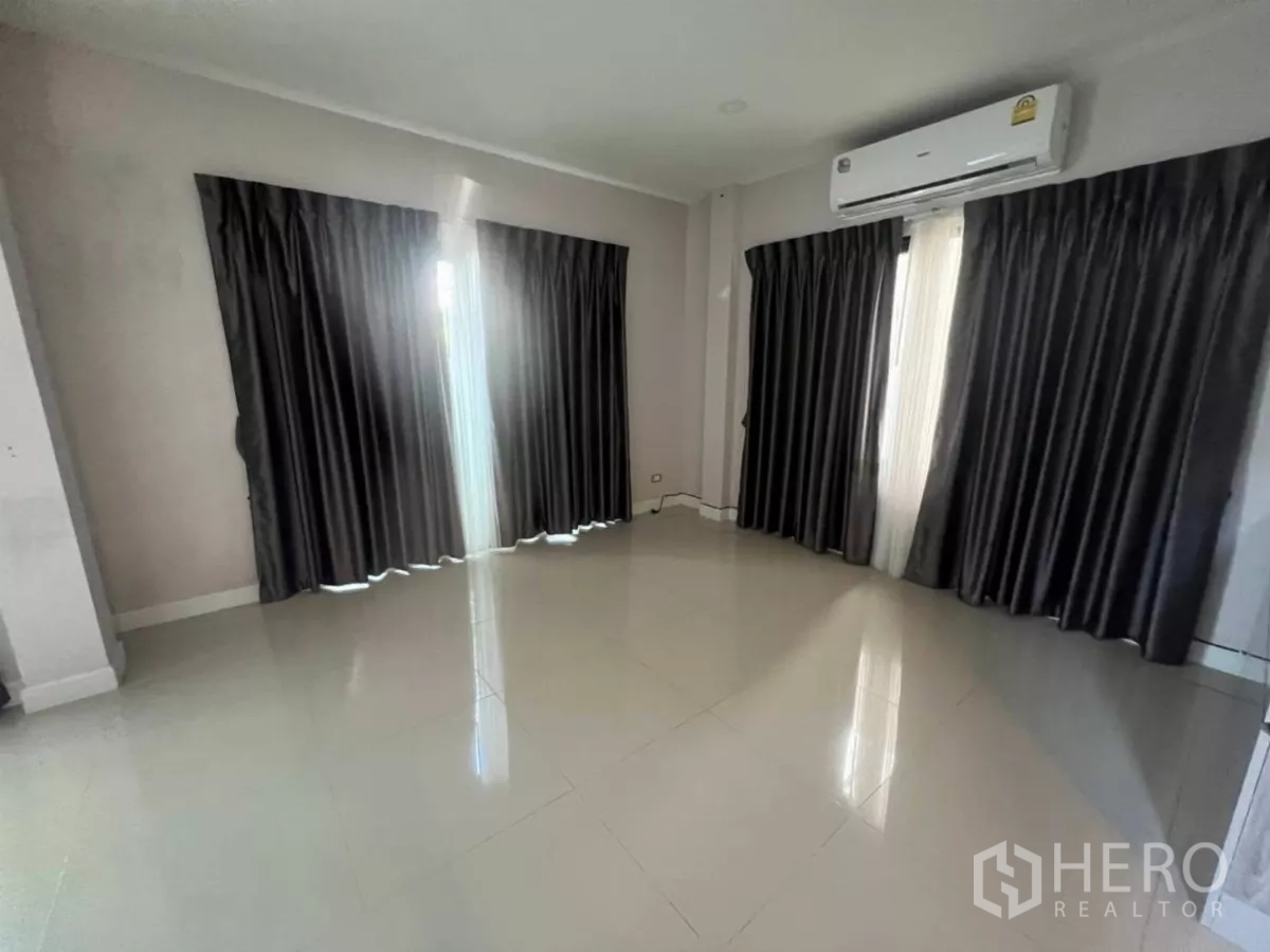 Detached House for rent in Samut Prakan - Bright empty living room with glossy tile floors, dark curtains and a wall-mounted air conditioner.